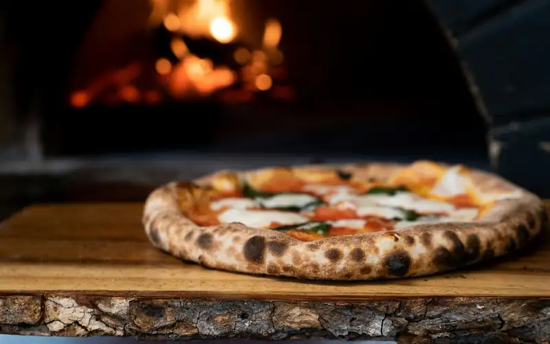 Napolitaanse pizza met geblakerde rand op een houten plank, met vuur op de achtergrond