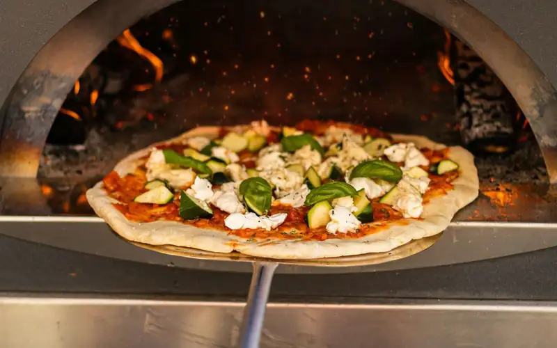 Pizza op een metalen schep voor de opening van een roestvrijstalen pizzaoven met vuur achterin